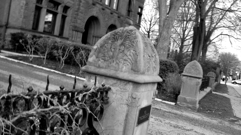 Carved stone gateposts and an iron fence before a sandstone building, in black and white.