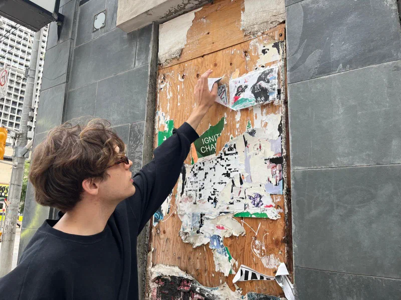 Andrada at a plywood hoarding layered with torn posters, Toronto, 2024.