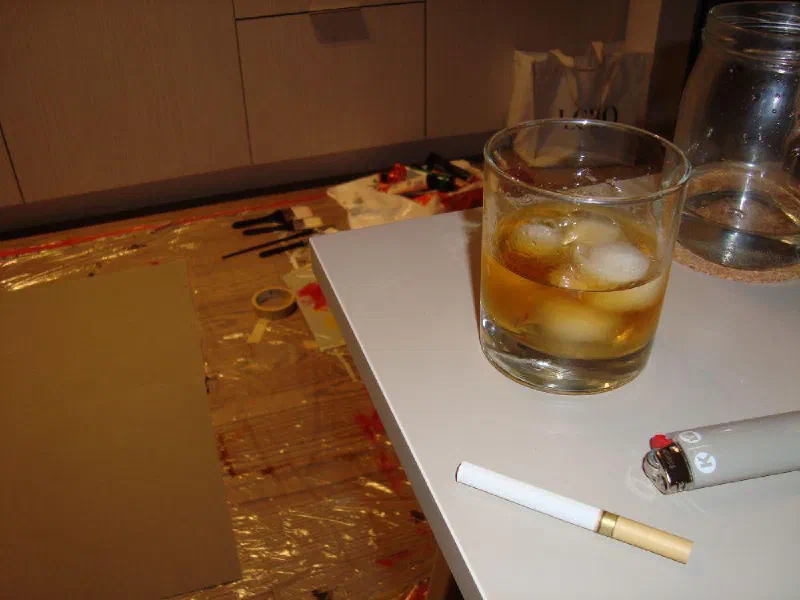 A glass of whisky and a cigarette beside a painting in progress on the floor of the first studio, 2024.