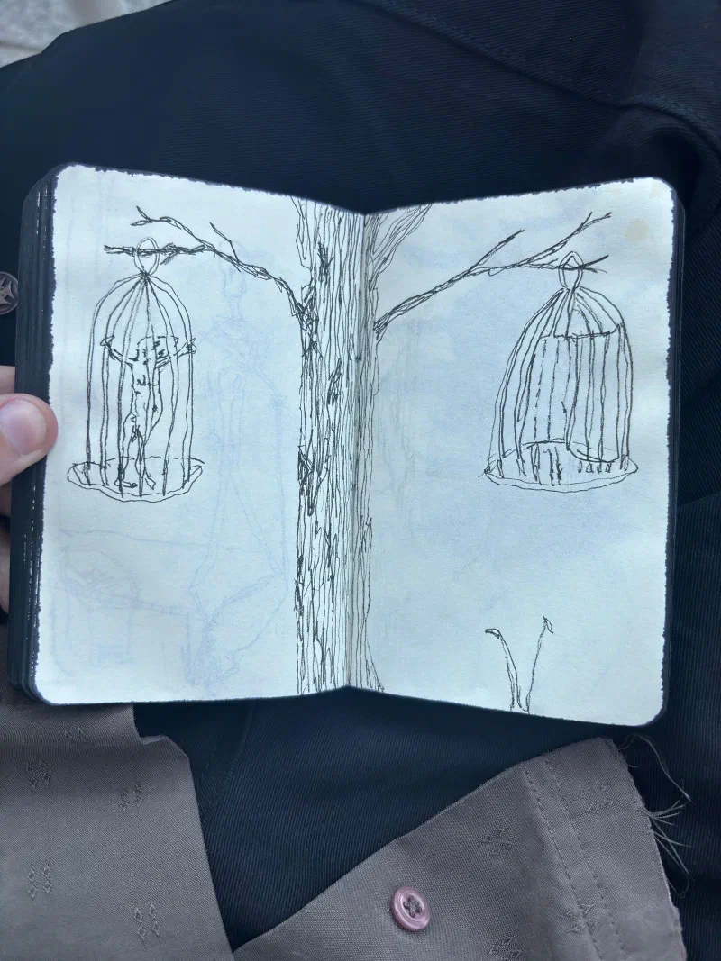 A sketchbook spread: two birdcages hanging from a tree, one holding a figure, July 2026.